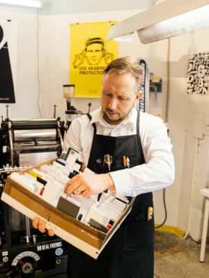 Adult man organizing printing supplies in a workshop with industrial machines around him.