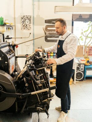 An adult male operates a vintage printing machine inside an industrial workshop setting.